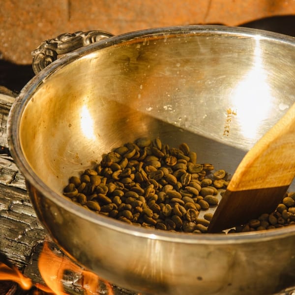 Coffee beans roasting in a metal pan with a wooden spoon over an open fire.