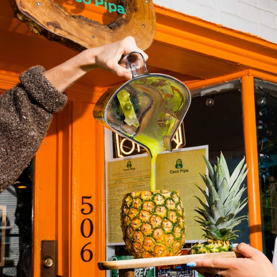 Green juice poured from a pitcher into a hollowed pineapple, outside a vibrant orange storefront.