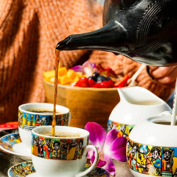 Coffee pouring from a black pot into a colorful cup, with fruit and cream nearby.