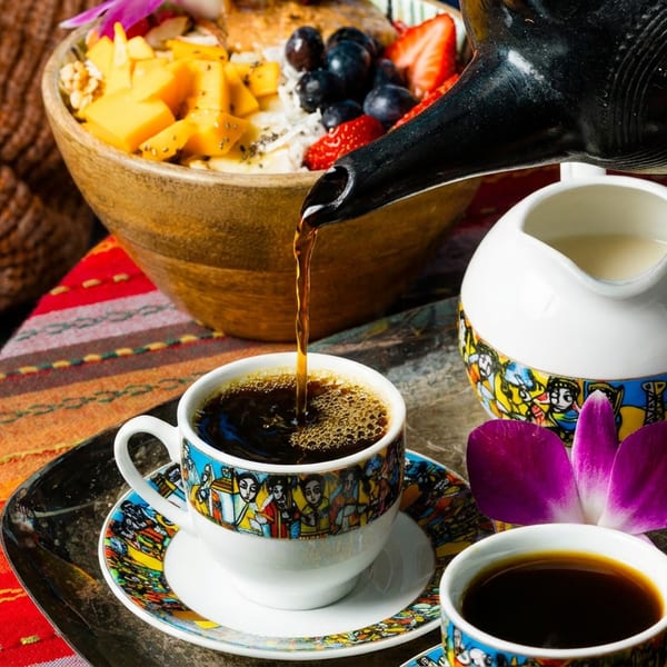 Coffee pouring into a colorful cup, with a fruit bowl and cream on a patterned tray.