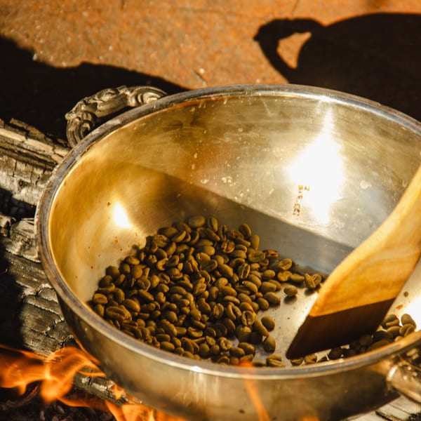 Green coffee beans roasting in a metal pan over an open fire with a wooden paddle.