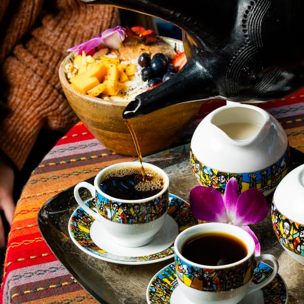 Coffee pouring from a black pot into a colorful cup, with a fruit bowl nearby.
