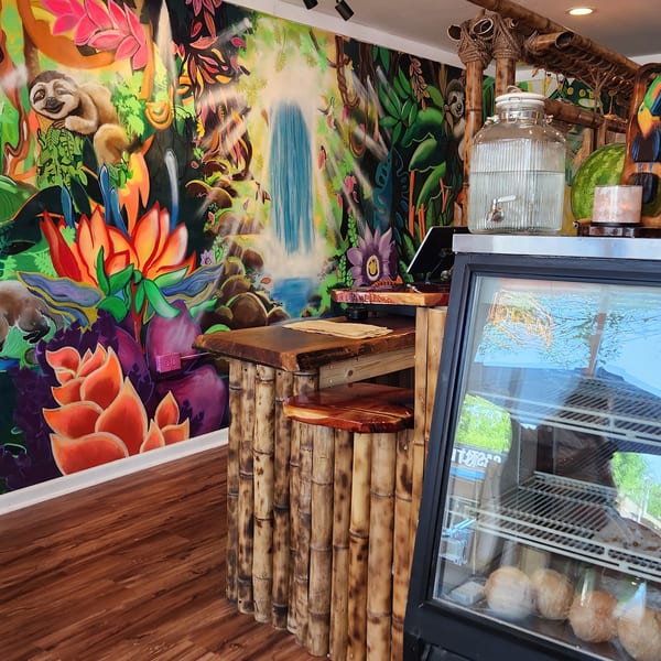A vibrant jungle mural with a waterfall, sloth, and colorful flowers behind a bamboo counter and display fridge.