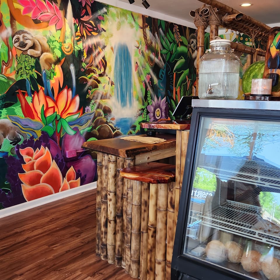 A vibrant jungle mural with a waterfall, sloth, and colorful flowers behind a bamboo counter and display fridge.