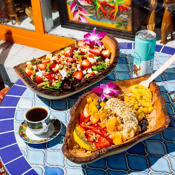 Two wooden bowls of fruit salad and green salad, coffee, and a drink on a blue tiled table.