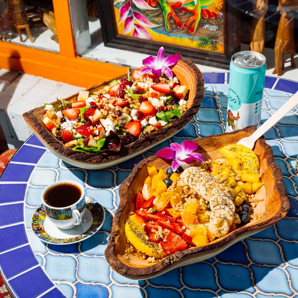 Two wooden bowls of fruit salad and green salad, coffee, and a drink on a blue tiled table.