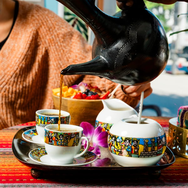 Coffee pouring from a traditional jebena into colorful cups on a tray with fruit and flowers.