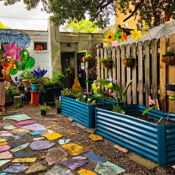 Two women stand in a vibrant community garden with colorful murals, raised beds, and a stone path.