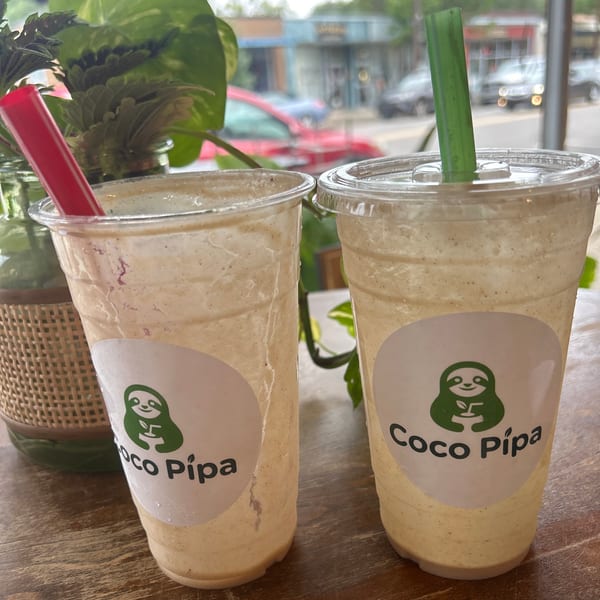 Two Coco Pipa smoothies with red and green straws on a wooden table, with a street view outside.